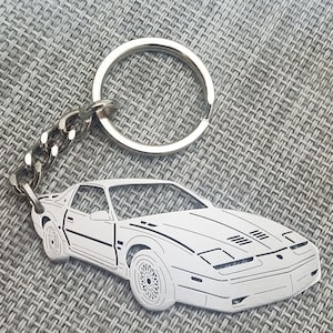May include: A silver keychain featuring a detailed outline of a white sports car. The car has a black stripe running along the side and a black grill with a honeycomb pattern.