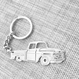 May include: Silver-colored keychain featuring a classic pickup truck design. The truck is detailed with headlights, a grille, and a license plate with the number 833429. The keychain includes a chain and a circular key ring.