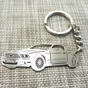 May include: A silver metal keychain featuring a detailed cutout of a Ford Mustang. The keychain has a silver ring and chain. The car has a license plate that reads "M1LRLTE".