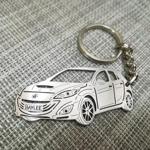 May include: A silver metal keychain featuring a detailed outline of a car with the word "BAYLEE" on the license plate. The keychain has a silver metal ring and chain.