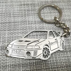 Puede incluir: Un llavero de metal plateado con un recorte detallado de un coche plateado con el texto "FT-G-37" en la parte delantera y "Georg" en el lateral. El llavero está unido a un anillo y una cadena plateados.