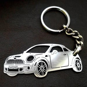 Puede incluir: Llavero de metal plateado con un recorte detallado de un coche Mini Cooper plateado. El llavero tiene un anillo y una cadena plateados.