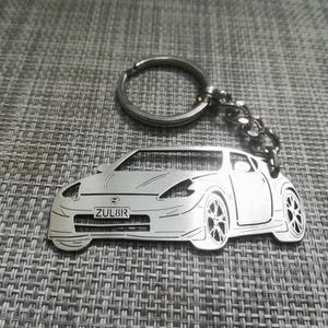 May include: A silver metal keychain featuring a detailed silhouette of a Nissan Z car. The keychain has a silver ring and chain. The car has a license plate that reads "ZUL8IR".