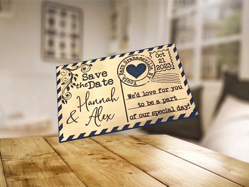 Pode incluir: Um cart&atilde;o de madeira dourado para guardar a data com uma borda listrada azul e branca. O cart&atilde;o tem um design de cart&atilde;o postal vintage com um selo em forma de cora&ccedil;&atilde;o que diz "You're Invited" e uma data de 21 de outubro de 2023. O texto do cart&atilde;o diz "Save the Date Hannah & Alex We'd love for you to be a part of our special day!"