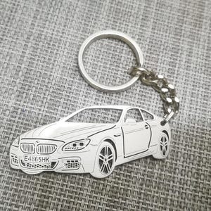 Pode incluir: Um chaveiro de prata com um contorno detalhado de um carro branco com a placa de matrícula "E486SHK".