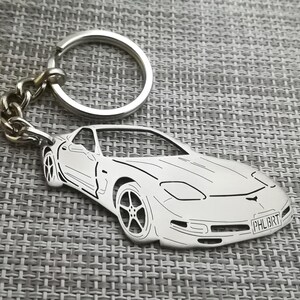 以下が含まれることがあります： 白いコルベットの輪郭を詳細に描いたシルバーのキーホルダー。ナンバープレートは「PHL8RT」。