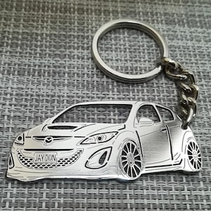 Könnte beinhalten: Ein silberner Schlüsselanhänger mit einer detaillierten Ausschneidung eines Autos mit dem Namen "JAYDON" auf dem Nummernschild. Der Schlüsselanhänger hat einen silbernen Ring und eine Kette.