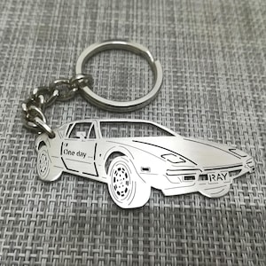 May include: A silver metal keychain featuring a detailed cutout of a sports car with the words "One day..." and "RAY" engraved on it.
