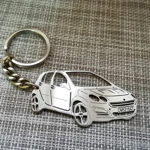 Op de afbeelding: Een zilveren metalen sleutelhanger met een gedetailleerde uitsnijding van een kleine auto met het kenteken "CM5315AT".