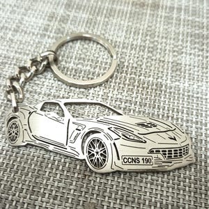 Puede incluir: Un llavero de metal plateado con un contorno detallado de un coche deportivo con el texto "CCNS 190" en la matrícula.