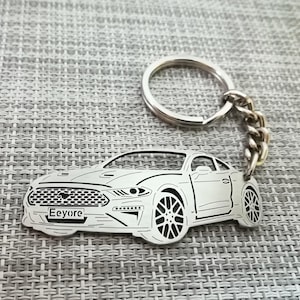 Puede incluir: Un llavero de metal plateado con un contorno detallado de un coche Ford Mustang. El llavero tiene un anillo y una cadena plateados. El coche tiene el nombre "Eeyore" grabado en la matrícula.