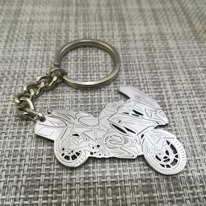 Puede incluir: Un llavero de metal plateado con un contorno detallado de una motocicleta. El llavero tiene un anillo de metal plateado para sujetarlo a las llaves.
