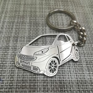 Op de afbeelding: Een zilveren metalen sleutelhanger met een gedetailleerde uitsnijding van een Smart-auto. De sleutelhanger heeft een zilveren ring en ketting.