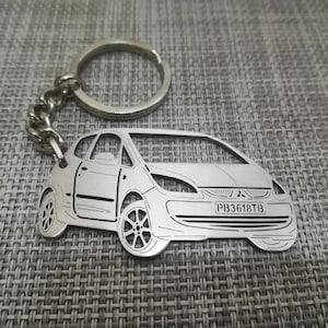 Könnte beinhalten: Ein silberner Schlüsselanhänger mit einer detaillierten Ausschneidung eines weißen Mitsubishi Colt Autos mit dem Kennzeichen "PB3618TB".