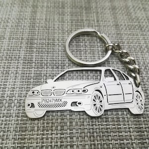 May include: Silver car-shaped keyring with a key ring and chain. The keyring depicts a detailed outline of a car, including headlights, wheels, and a license plate with the text "PI32479MX". The background is a textured grey surface.