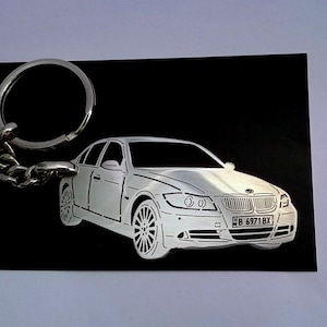 May include: A silver-colored car keychain with a detailed car outline on a black background. The keychain includes a key ring and chain. The car design features headlights, wheels, and a license plate.