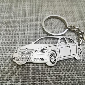 Op de afbeelding: Een zilveren metalen sleutelhanger met een gedetailleerde omtrek van een witte auto met een kentekenplaat die "BP6935CC" aangeeft.