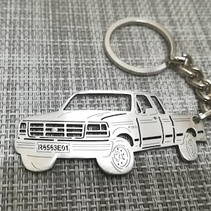 Può includere: Portachiavi in metallo argentato con un ritaglio dettagliato di un pick-up Ford F150. Il camion ha una targa che recita "R5553E01".