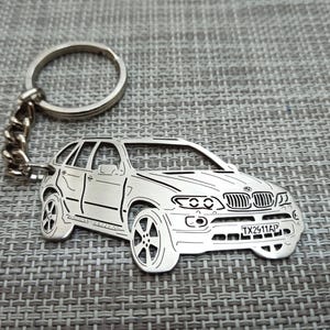 May include: Silver metal keychain featuring a detailed cutout of a silver BMW X5 SUV with a license plate that reads "TX2511AP".