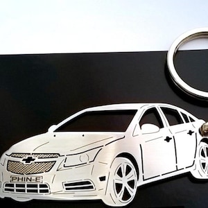 May include: A silver metal keychain featuring a cutout design of a white Chevrolet car. The keychain has a silver ring and a silver clasp. The car has the text "PHIN-E" on the front.