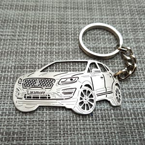 May include: A silver metal keychain featuring a detailed outline of a black Lincoln SUV. The keychain has a silver ring and chain. The car has the word "Laramore" engraved on the front.