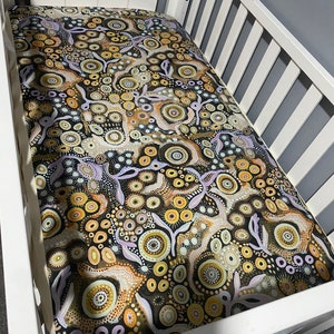 May include: A white wooden cot frame fitted with a mattress cover featuring a complex, dense pattern of concentric circles, dots, and organic shapes in a colour palette of black, mustard yellow, tan, white, and soft lavender throughout the fabric.