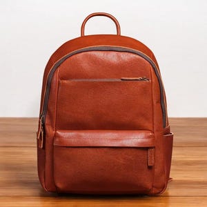 K&ouml;nnte beinhalten: Ein brauner Lederrucksack mit einer Fronttasche und einem Rei&szlig;verschluss. Der Rucksack hat einen Tragegriff oben und verstellbare Schultergurte.