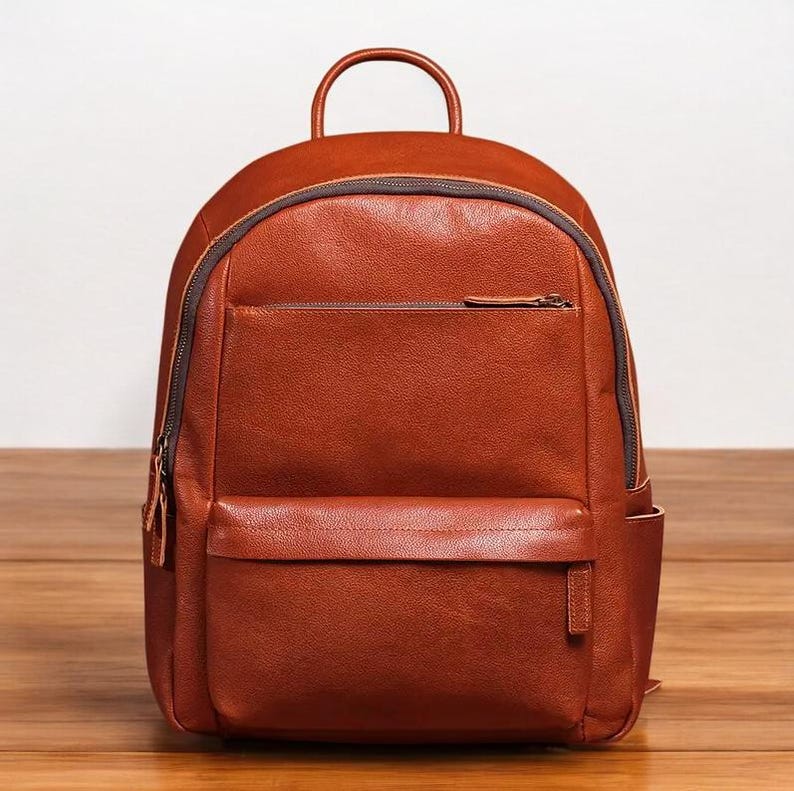 K&ouml;nnte beinhalten: Ein brauner Lederrucksack mit einer Fronttasche und einem Rei&szlig;verschluss. Der Rucksack hat einen Tragegriff oben und verstellbare Schultergurte.
