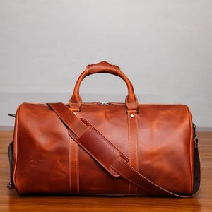 K&ouml;nnte beinhalten: Eine braune Leder-Reisetasche mit einem oberen Griff und einem Schultergurt. Die Tasche hat einen Rei&szlig;verschluss und eine Fronttasche.
