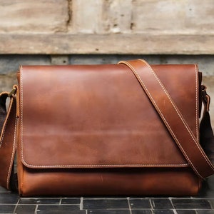 K&ouml;nnte beinhalten: Eine braune Ledertasche mit einem langen, verstellbaren Trageriemen. Die Tasche hat einen Klappverschluss und ein einzelnes Fach.