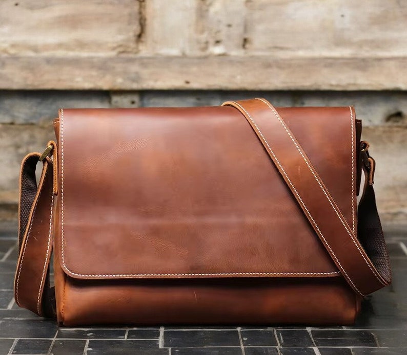 K&ouml;nnte beinhalten: Eine braune Ledertasche mit einem langen, verstellbaren Trageriemen. Die Tasche hat einen Klappverschluss und ein einzelnes Fach.