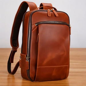 K&ouml;nnte beinhalten: Ein brauner Lederrucksack mit einer Rei&szlig;verschlusstasche vorne und verstellbaren Tr&auml;gern.