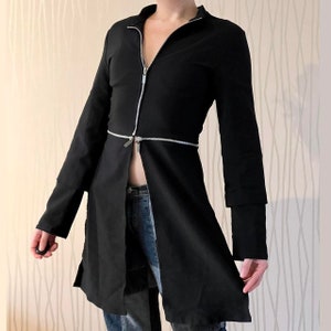 Puede incluir: Abrigo largo negro con cierre de cremallera plateada en la parte delantera y en la cintura. El abrigo tiene mangas largas con puños en capas y un diseño frontal dividido. El abrigo se usa con vaqueros azules.