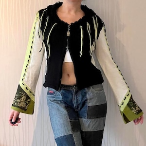 Könnte beinhalten: Eine schwarze und weiße Strickjacke mit grünem Fransenbesatz und Reißverschluss. Die Jacke hat ein weiß-grünes Muster an den Ärmeln.