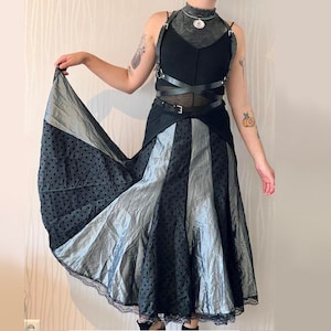 May include: A black and silver skirt with a polka dot pattern and a lace trim. The skirt is layered and has a flowing silhouette. The model is wearing a black top with a leather harness.