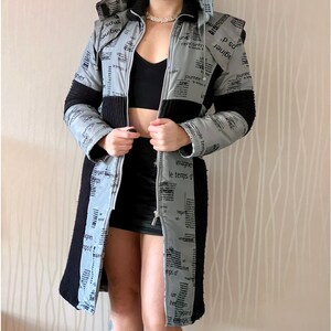 May include: A long gray and black hooded jacket with a zipper closure. The jacket is made of a quilted fabric with a newspaper print design. The text on the jacket includes the words "journee", "temps", "un", "imagine", and "regard".