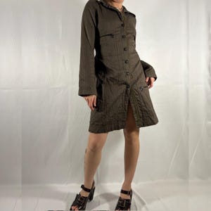 May include: A brown long-sleeved collared shirt dress with a button-up front and pockets. The dress has a slightly A-line silhouette and is made of a textured fabric. The model is wearing the dress with brown heeled sandals.