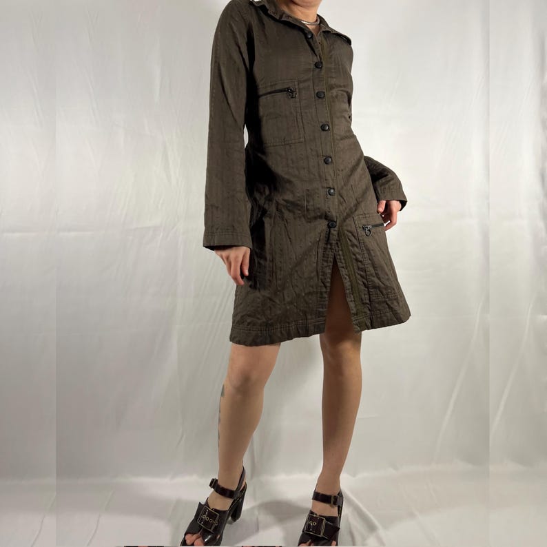May include: A brown long-sleeved collared shirt dress with a button-up front and pockets. The dress has a slightly A-line silhouette and is made of a textured fabric. The model is wearing the dress with brown heeled sandals.