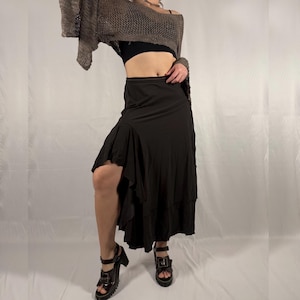 Puede incluir: Una falda asimétrica marrón con un dobladillo con volantes. La falda se combina con un top negro y sandalias de plataforma. Un top de punto suelto marrón de manga larga está drapeado sobre los hombros. El conjunto está sobre un fondo blanco.