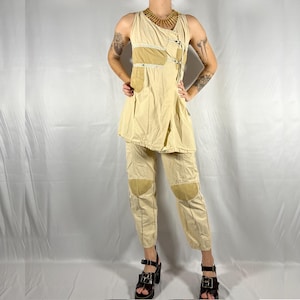 Puede incluir: Una blusa beige sin mangas con un diseño en capas y un pantalón beige a juego con parches en las rodillas. El atuendo se complementa con un collar de cuentas marrones.