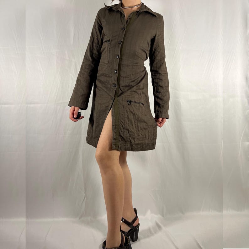 May include: A brown linen button-down shirt dress with long sleeves and a single front pocket. The dress has a slit up the front and is cinched at the waist.