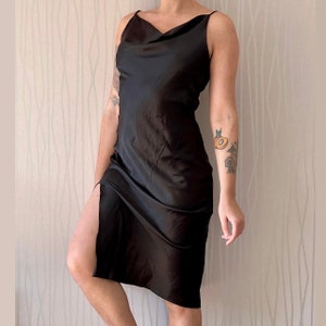 May include: A black satin slip dress with a cowl neck and a side slit. The dress is sleeveless and has a relaxed fit.