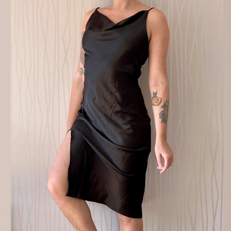 May include: A black satin slip dress with a cowl neck and a side slit. The dress is sleeveless and has a relaxed fit.