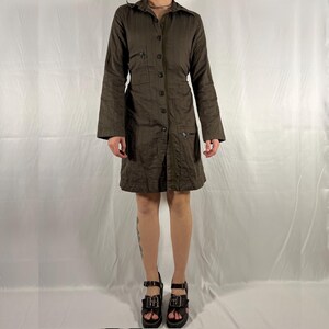May include: A brown, long-sleeved, button-up dress with a collared neckline and a fitted waist. The dress has multiple pockets and a zipper detail on the front. The model is wearing the dress with black platform sandals.