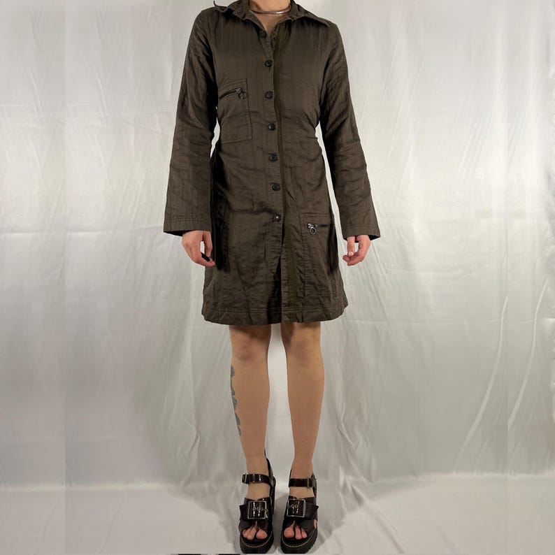 May include: A brown, long-sleeved, button-up dress with a collared neckline and a fitted waist. The dress has multiple pockets and a zipper detail on the front. The model is wearing the dress with black platform sandals.