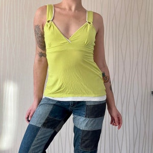 Puede incluir: Un top verde lima sin mangas con ribete blanco y cuello en V. El top tiene dos anillos metálicos en las tiras. La modelo lleva el top con unos vaqueros de mezclilla con parches.