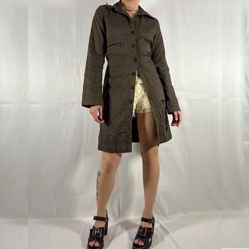 May include: A brown, long-sleeved shirt dress with a button-up front and a cinched waist. The dress has a relaxed fit and a slightly flared skirt. The model is wearing the dress with a pair of white lace shorts and brown heeled sandals.