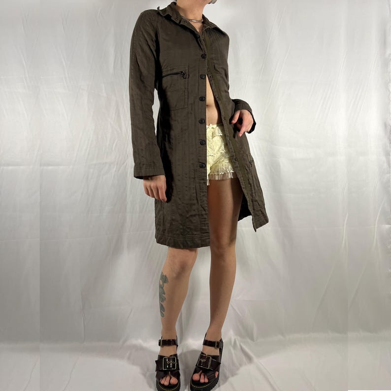 May include: A long brown button-down shirt with a relaxed fit. The shirt has a collar, long sleeves, and a button-up front. The model is wearing the shirt open, revealing a white lace undershirt.
