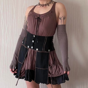 Pode incluir: Conjunto de top halter marrom e saia em camadas. A saia apresenta painéis pretos com detalhes quadrados e bainha com babados. Aquecedores de braço marrons combinando e colar de prata completam o traje. O estilo é boêmio e alternativo.