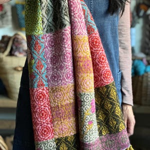 May include: A colorful patchwork scarf with a variety of geometric and floral patterns. The scarf features squares of green, pink, red, and yellow, with a yellow tassel fringe. The scarf is made of a soft, woven material.